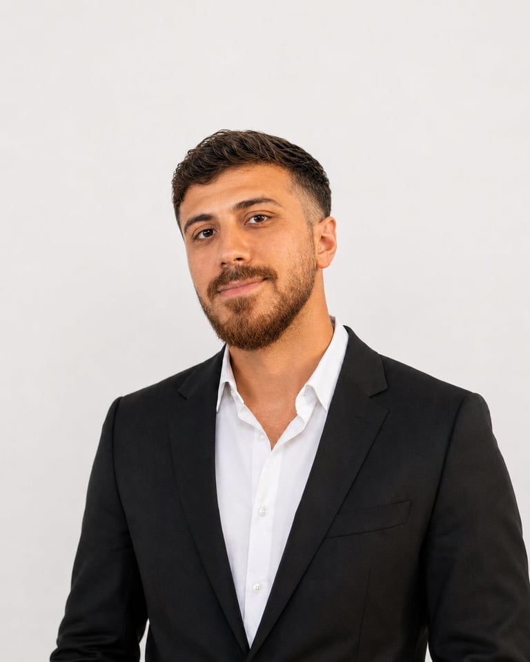 Professional headshot of a man with dark hair and beard wearing a black blazer over a white dress shirt against a light gray background.